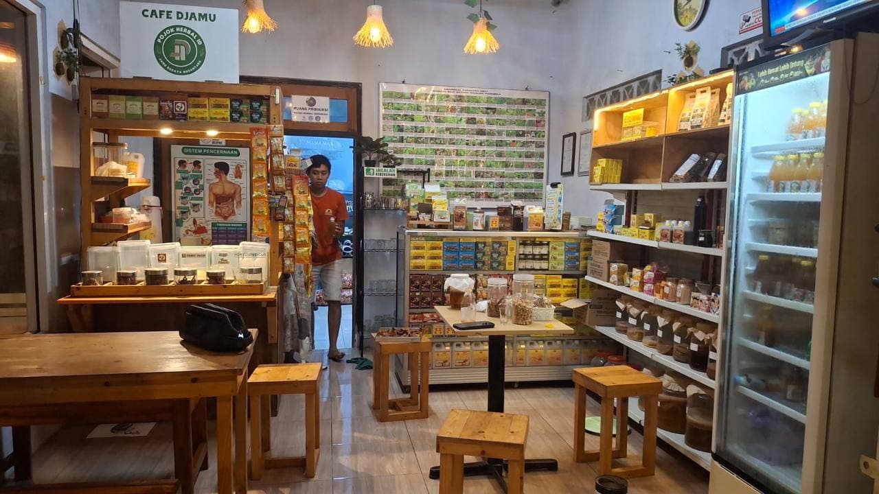 Interior Cafe Djamu Pojok Herbal: bangku dan meja kayu, pencahayaan hangat, etalase rempah dan kulkas botol jamu di dinding belakang