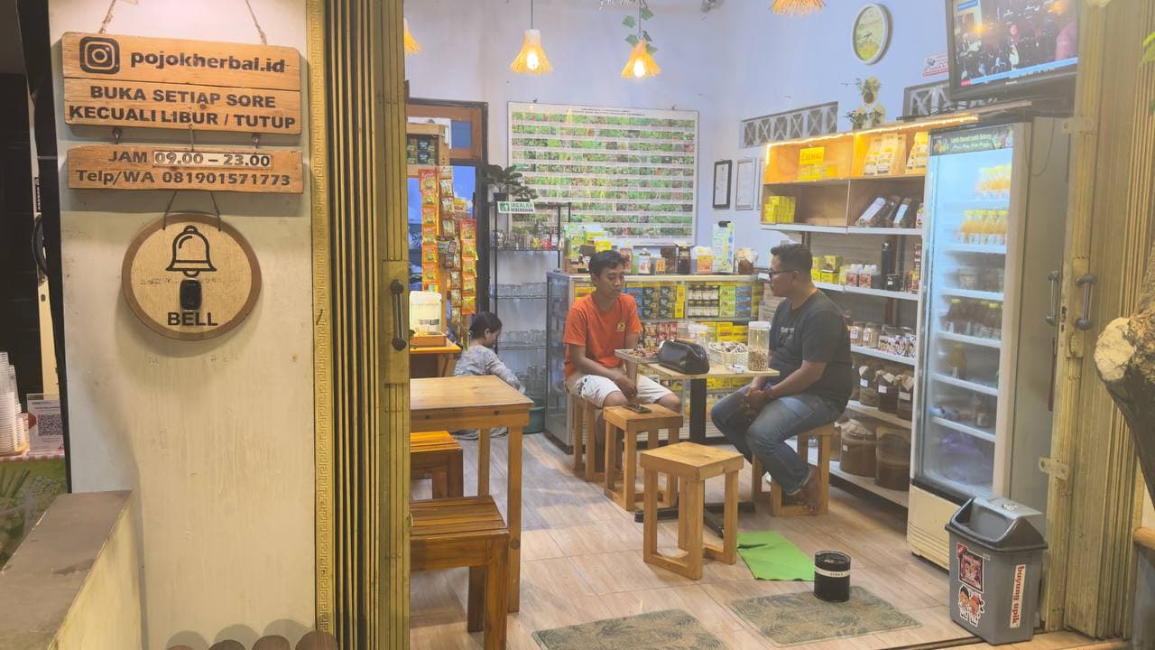 Tampak depan kedai Pojok Herbal — papan nama @pojokherbal.id dengan jam buka, anak muda berbincang santai di dalam kedai yang dipenuhi rak rempah dan etalase produk herbal